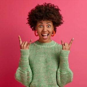 Happy carefree dark skinned rebellious young woman enjoys awesome music makes rock n roll gesture has fun on music festival or cool event wears casual jumper poses against pink wall home